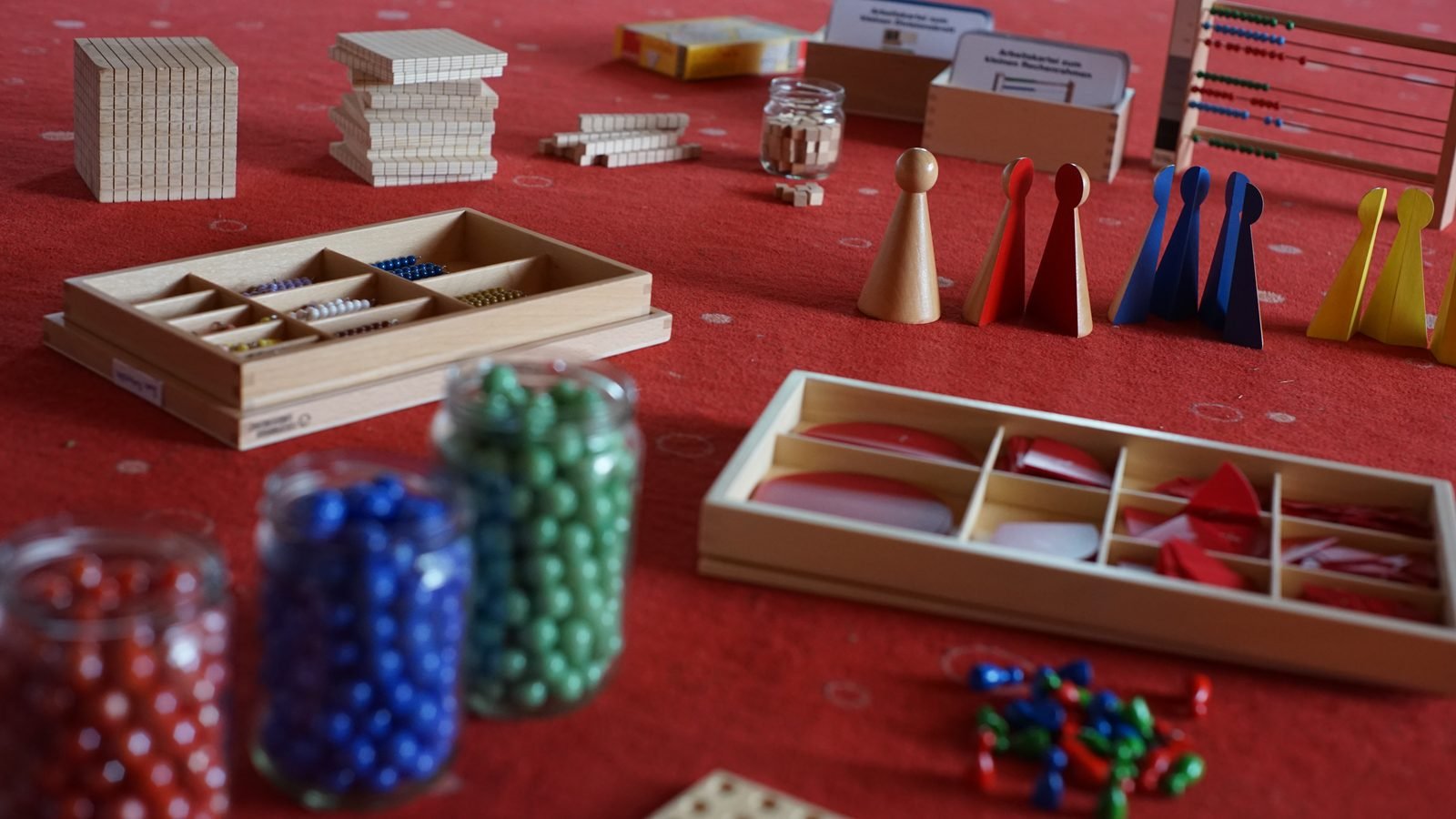 Montessori-Mathematikmaterial auf rotem Teppich: Goldenes Perlenmaterial, Holzwürfel und -stangen, Perlenketten, Rechenrahmen und Spielfiguren.